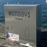 A white free-vend dispenser with the Mondays Organic brand visible, placed on a wooden surface with a body of water and sky in the background.