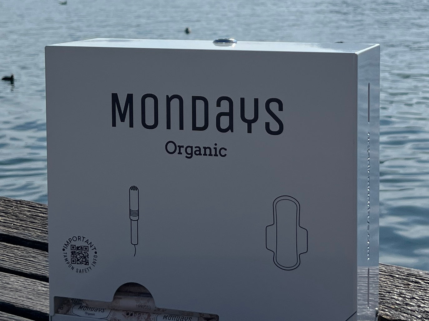 A box of Mondays Organic menstrual products, featuring the product name and an illustration of a tampon, placed on a wooden surface with a body of water and a mountain in the background.