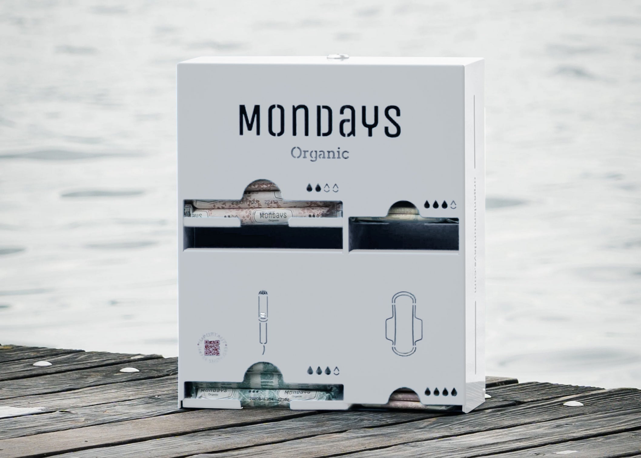 A stainless steel menstrual product dispenser in white with the brand name 'Mondays Organic' on the front, placed on a wooden surface with water in the background.