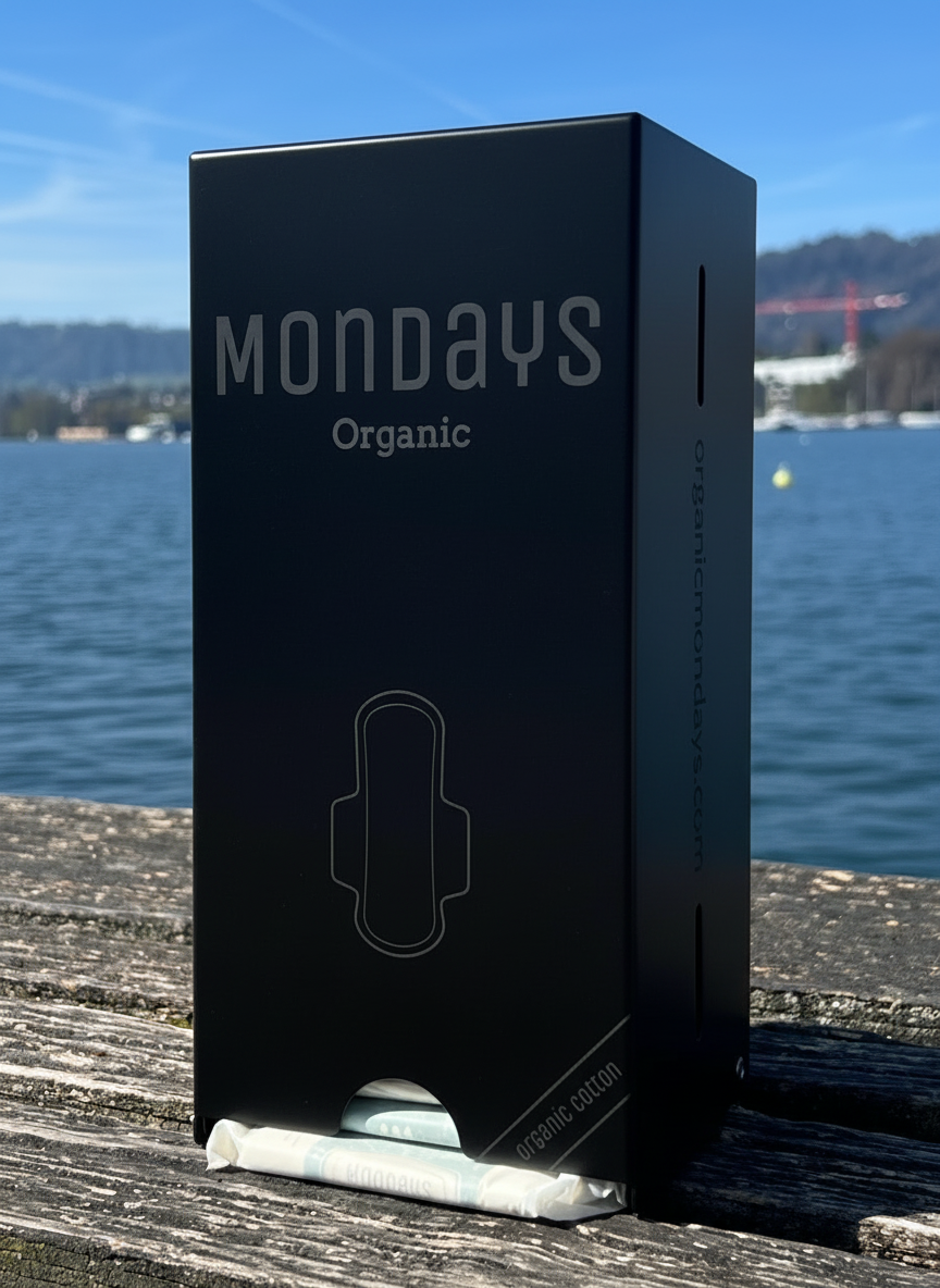 Black pad dispenser ideal for schools, venues and sportsclubs providing an essential sanitary product for 50% of population. On a wooden surface with a scenic background.