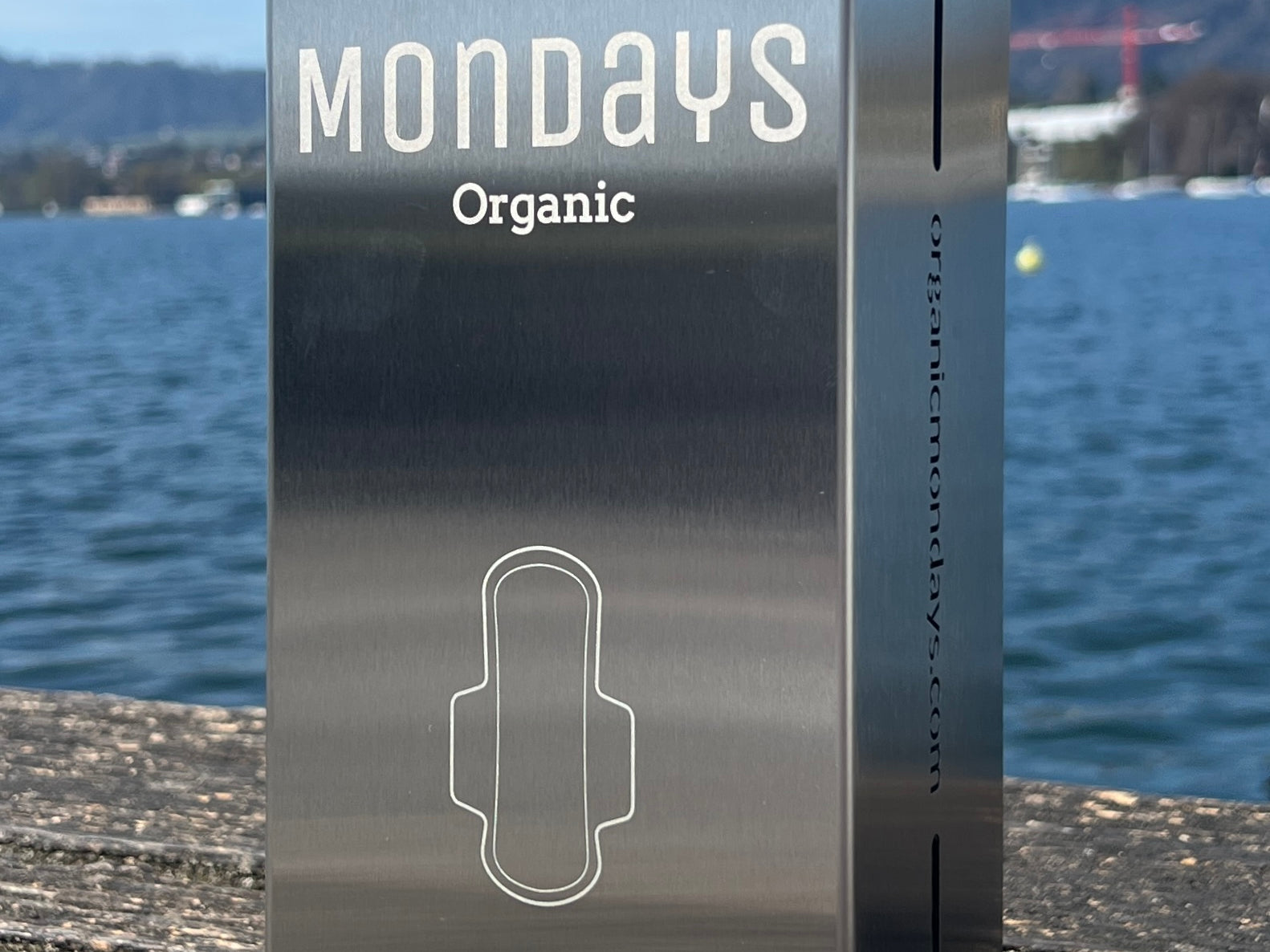 A stainless steel compact pad dispenser with the Mondays Organic brand visible, placed on a wooden surface with a body of water and a hilly landscape in the background.