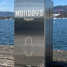 A stainless steel compact pad dispenser with the Mondays Organic brand visible, placed on a wooden surface with a body of water and a hilly landscape in the background.