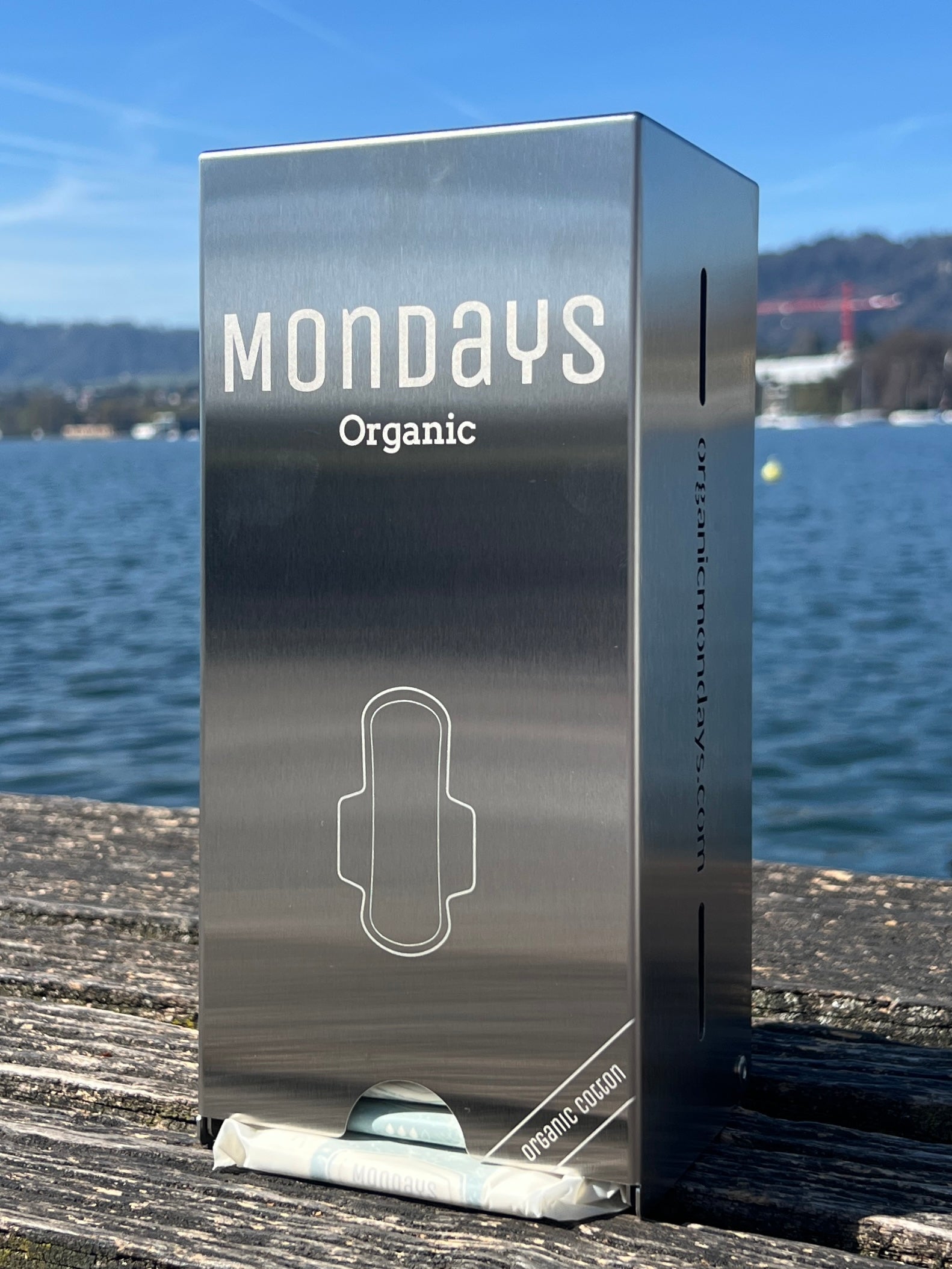 A stainless steel compact pad dispenser with the Mondays Organic brand visible, placed on a wooden surface with a body of water and a hilly landscape in the background.