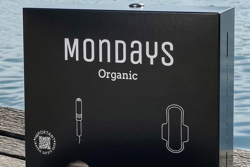 Mondays Organic BLACK dispenser for organic sanitary pads and applicator tampons sitting on a wooden surface with a lake and mountains in the background
