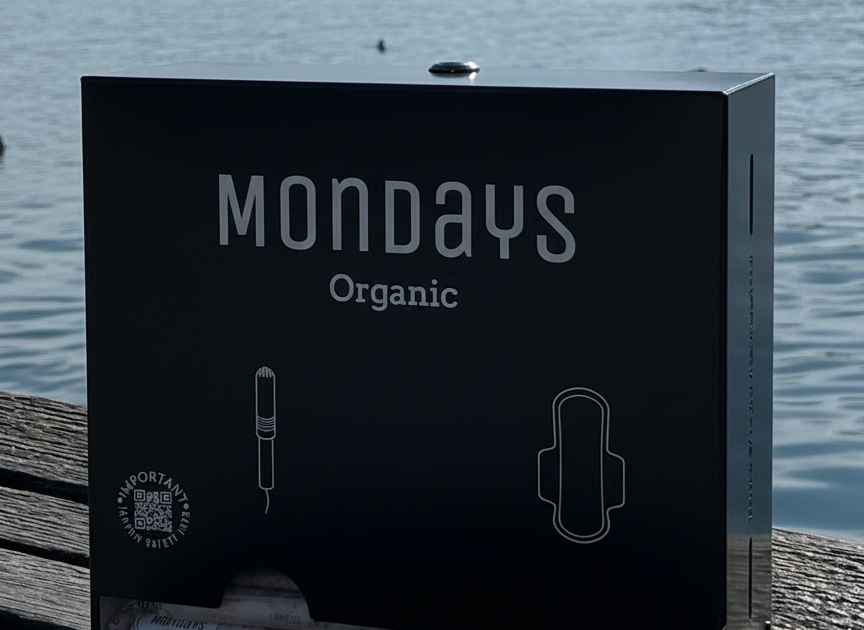 Mondays Organic BLACK dispenser for organic sanitary pads and applicator tampons sitting on a wooden surface with a lake and mountains in the background
