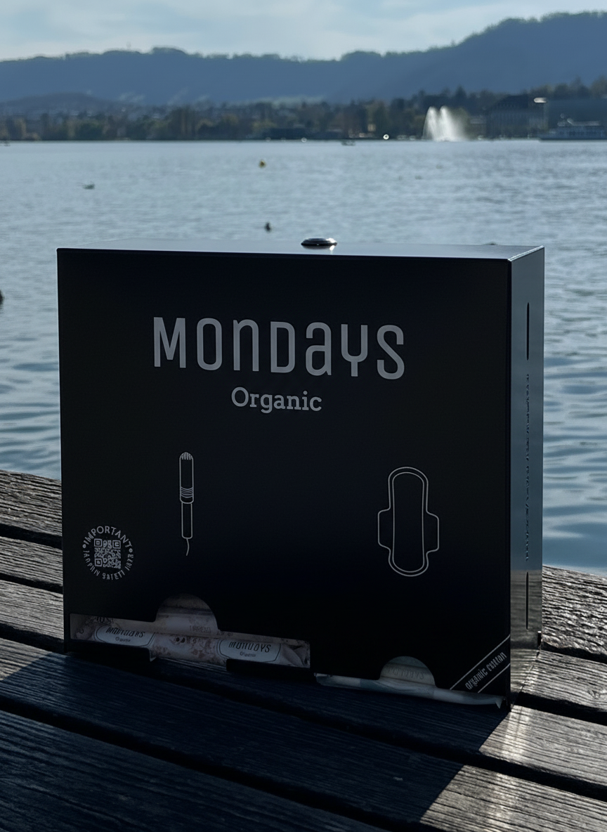 Mondays Organic BLACK dispenser for organic sanitary pads and applicator tampons sitting on a wooden surface with a lake and mountains in the background