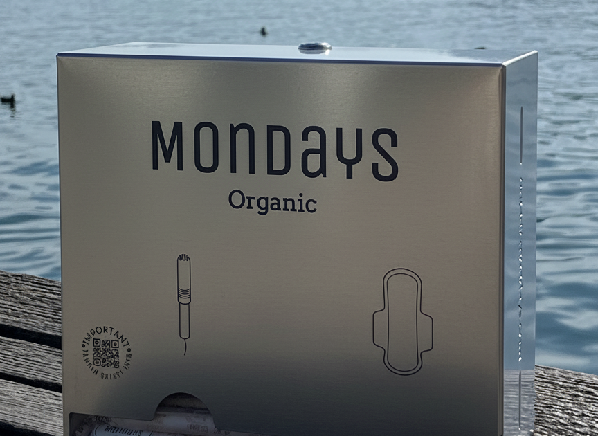Mondays Organic pad and tampon dispenser made of stainless steel stiting on a wooden surface with a lake and mountains in the background. Elegant, compact and durable. An investment in your people that pays back time after time.