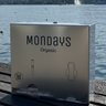Mondays Organic pad and tampon dispenser made of stainless steel stiting on a wooden surface with a lake and mountains in the background. Elegant, compact and durable. An investment in your people that pays back time after time.