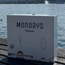 A box of Mondays Organic menstrual products, featuring the product name and an illustration of a tampon, placed on a wooden surface with a body of water and a mountain in the background.