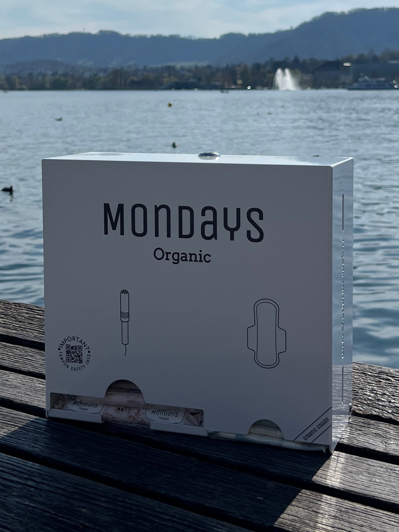 A box of Mondays Organic menstrual products, featuring the product name and an illustration of a tampon, placed on a wooden surface with a body of water and a mountain in the background.