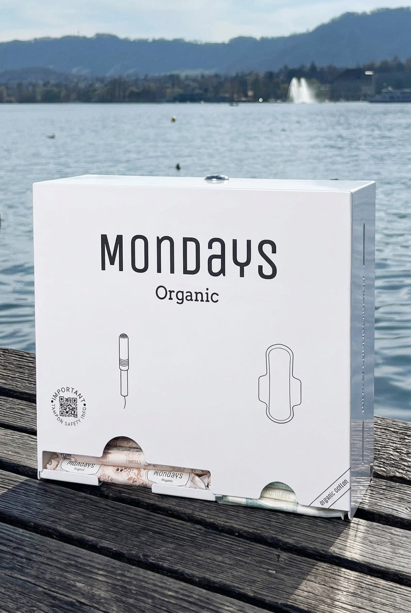 A box of Mondays Organic menstrual products, featuring the product name and an illustration of a tampon, placed on a wooden surface with a body of water and a mountain in the background.