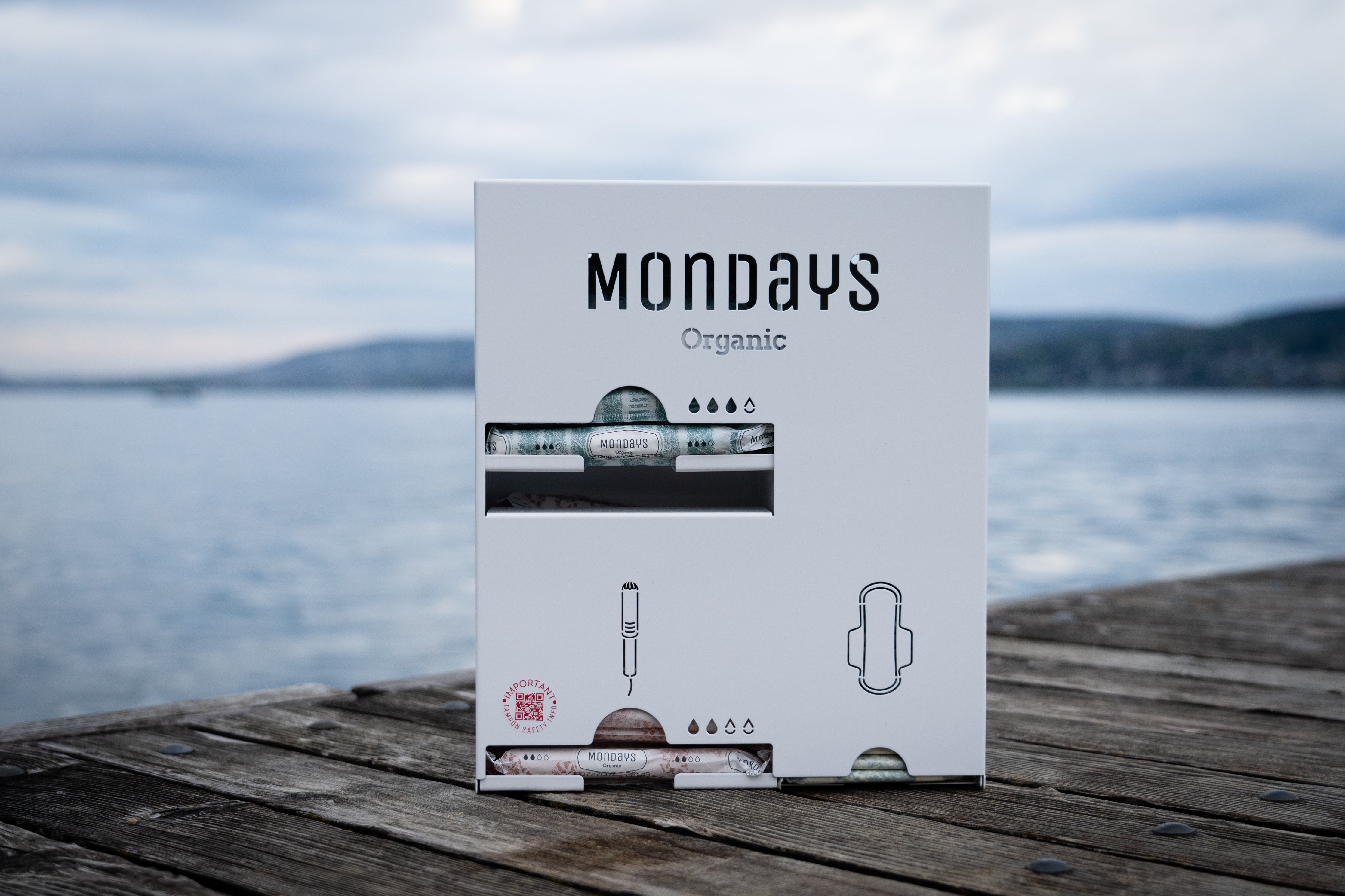A Mondays brand menstrual hygiene product dispenser placed on a wooden surface with a body of water and a hilly landscape in the background.